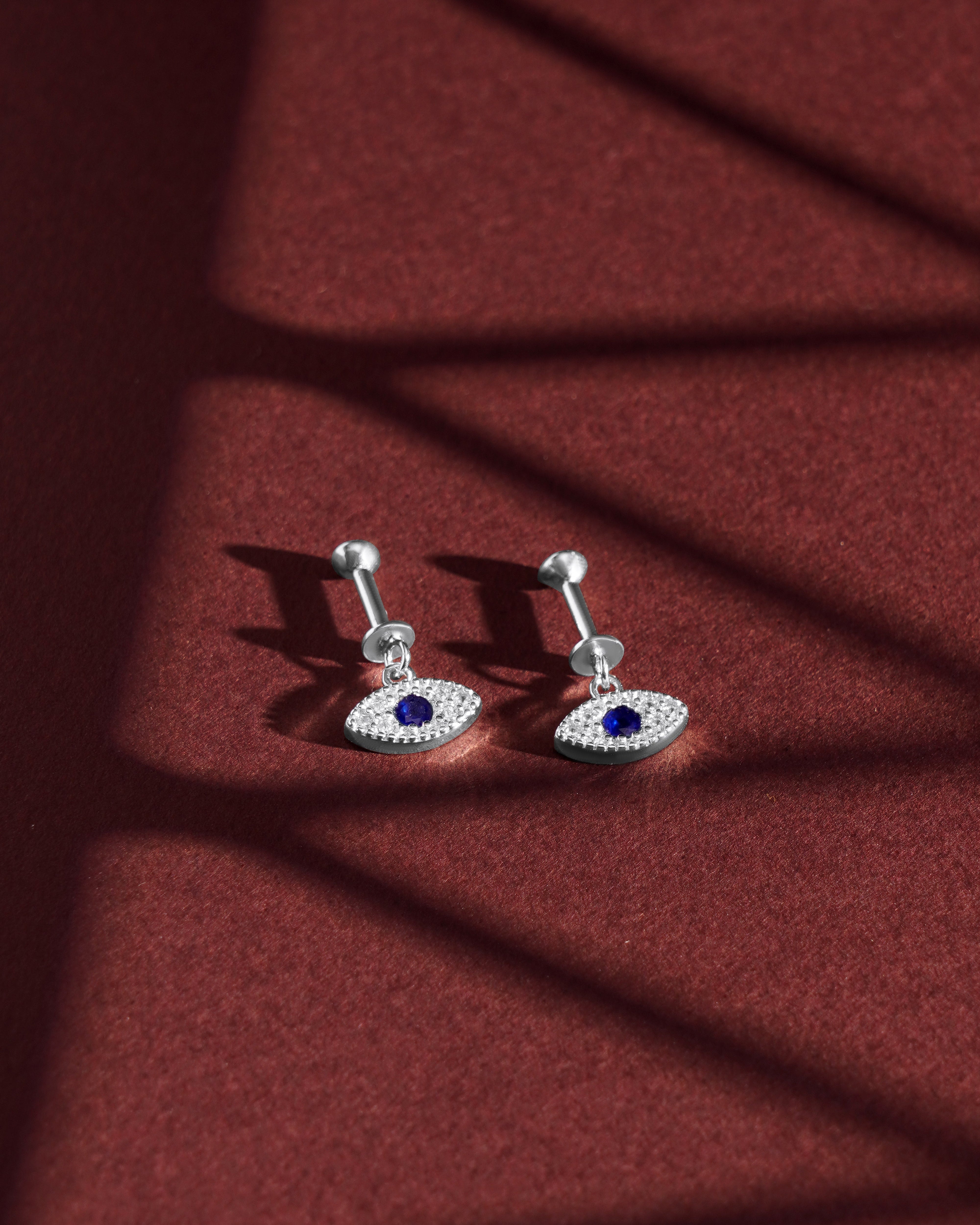 Zirconia Small Evil Eye Earrings - Versatile Style | Hanging Pair displayed on a red textured surface with dramatic shadows, showcasing their sparkle.